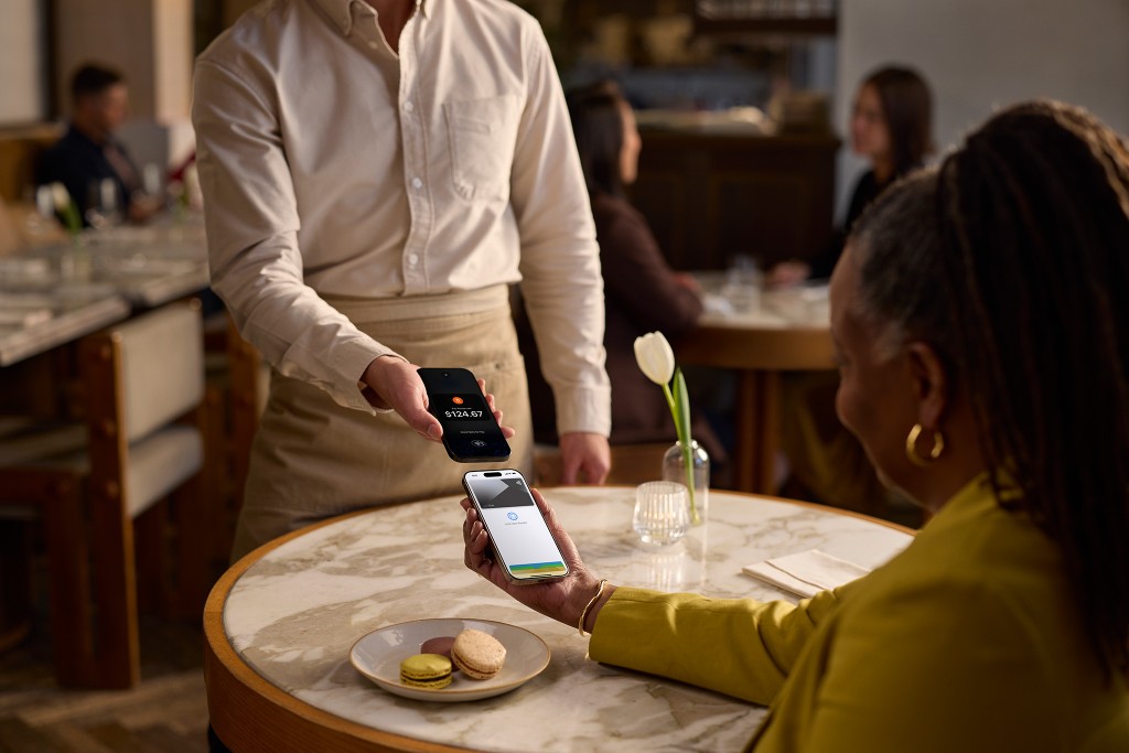 A server holds an iPhone showing a $124.67 charge while a seated diner pays with their own iPhone.
